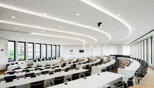 Auch die gestalterischen Fähigkeiten der Gipskartondecke kommen im großen Sitzungsaal eindrucksvoll zum Tragen: So wird der stufenweise Anstieg der Sitzreihen durch den treppenartigen Verlauf der Klimadecke eindrucksvoll aufgegriffen. Bildquelle: QLIMATE GmbH, Lahr